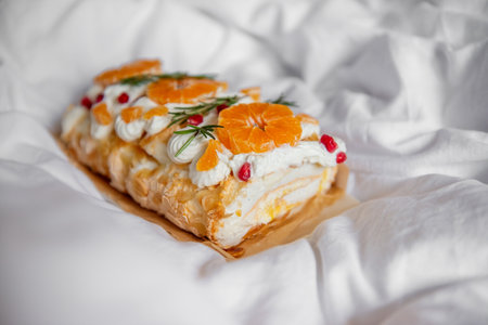 A close-up of a decorative pavlova roll topped with fresh orange slices, pomegranate seeds, and whipped cream, presented on a white background. Festive sweet dessertの写真素材