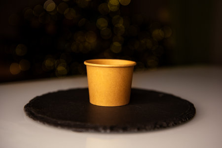 Small Craft paper cup sits on a black slate plate with warm golden bokeh lights in the background, creating a cozy and festive ambiance.の写真素材