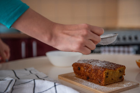 Confectioner sprinkles powdered sugar over a freshly baked loaf cake on a wooden board in a cozy...