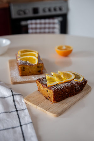 Two freshly baked orange cakes topped with orange slices and powdered sugar sit on wooden boards in a cozy kitchen, ready to be served.の写真素材