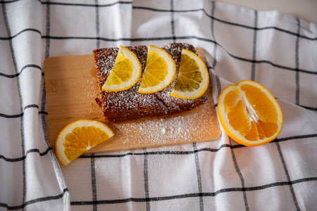 Freshly baked orange cake garnished with orange slices and dusted with powdered sugar, placed on a wooden board over a checkered cloth for a cozy, homemade touch.の写真素材
