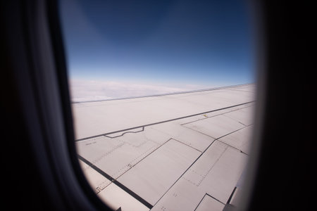 View from an airplane window showing the wing during a flight, with clouds below and a clear blue...
