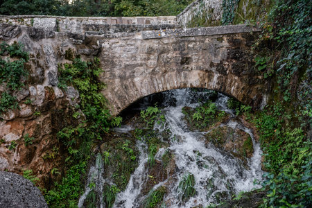 Water flowing beneath an ancient stone bridge cascades over moss-covered rocks, creating a serene...