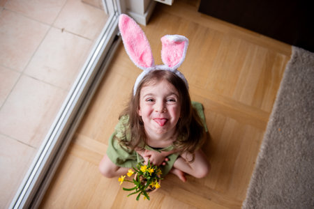 Top view of Playful little girl is sitting on the floor, wearing bunny ears and holding a pot of yellow flowers, sticking her tongue out in a funny expressionの写真素材