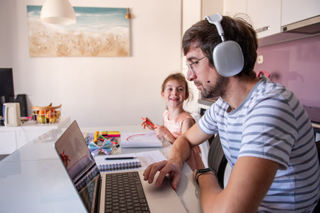 Father working from home, wearing headphones, while his little daughter happily drawing beside him at the kitchen table, creating a warm and supportive family atmosphereの写真素材