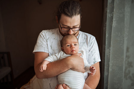 Father lovingly holds his baby near a sunlit window, creating a serene and intimate moment. The soft...