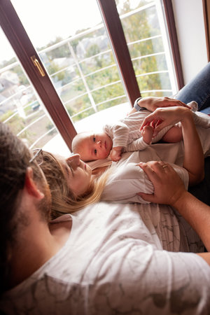 Young parents lying on the windowsill, cuddling and embracing their newborn baby, are creating a...