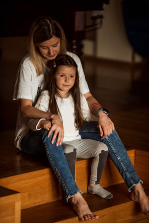 Nurturing mother lovingly embraces her young daughter as they sit together on wooden steps, enjoying a quiet and intimate moment. The warm indoor setting enhances their connectionの写真素材
