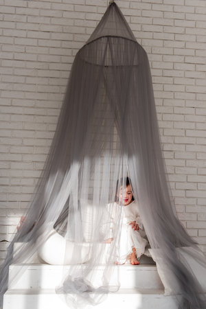 Young child sits contemplatively inside a mosquito net draped over a small set of stairs in a modern, minimalist bedroom, enjoying a quiet moment of relaxationの写真素材