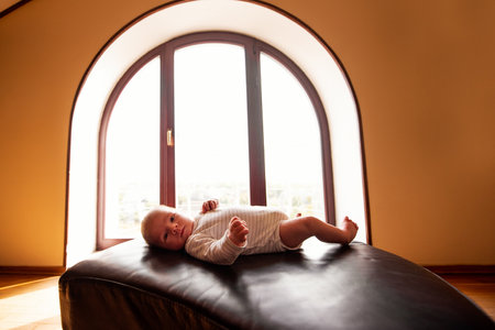 Caucasian baby lying comfortably on a leather sofa, basking in warm sunlight streaming through a large arched window in a modern apartment, creating a serene and cozy atmosphereの写真素材
