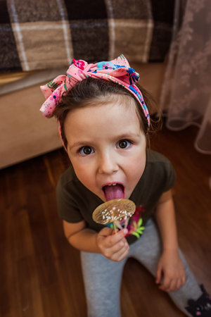 Young girl with a floral headband happily savoring a large, decorative lollipop while sitting on a wooden floor. The cozy setting includes soft furnishings and natural lightingの写真素材