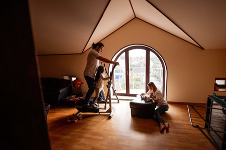 Father and daughter exercising on an elliptical machine while mother caring for their newborn baby in the living room, creating a lively and healthy family atmosphere at homeの写真素材