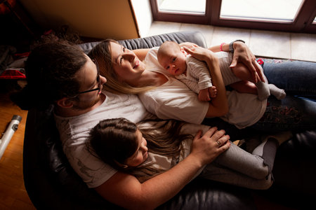 Parents lying on a cozy sofa with their newborn baby and daughter, savoring a peaceful moment together at home, embodying love and togetherness in a serene atmosphereの写真素材