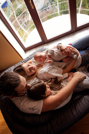 Family of four, including a newborn, enjoys a peaceful moment together on a comfortable sofa near a large window, embracing the warmth and love of their home environmentの写真素材
