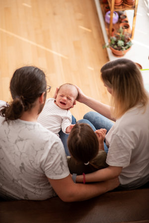 Parents comforting their crying newborn while sitting on the floor, holding their toddler daughter close. Embracing moments of love and support in a warm, nurturing home environmentの写真素材