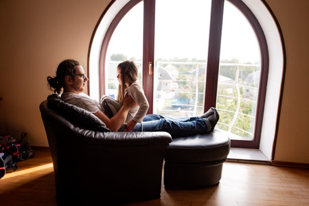 Father is sitting on a comfortable armchair with his feet up on a footstool while holding his daughter and looking out a large arched window in their modern homeの写真素材