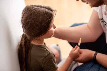 Dad giving a lollipop to his little daughter, sharing a sweet moment of family life at home, promoting love, care, and connection between parent and childの写真素材