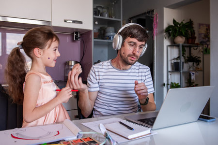 Father wearing headphones and working from home at kitchen table having video call on laptop, gesturing with hands while his little daughter drawing next to himの写真素材