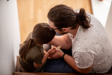 Loving father comforting his sad little daughter while sitting on stairs at home, supporting each other during difficult times, parenting and family support conceptの写真素材