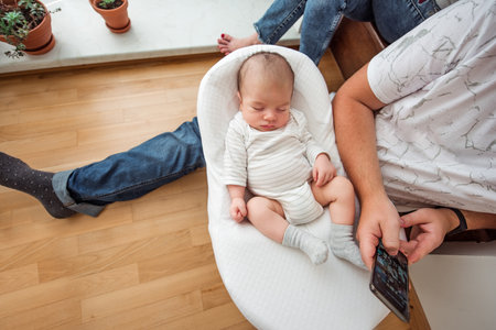 Peaceful scene of a baby sleeping in a cozy infant seat while a parent sits nearby using a smartphone. The setting includes wooden floors and potted plants, creating a warm, homely atmosphereの写真素材