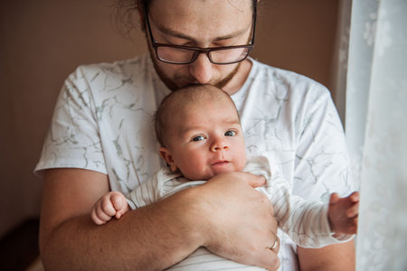 Tender moment as a father cradles his newborn baby near a window. The soft lighting highlights the...