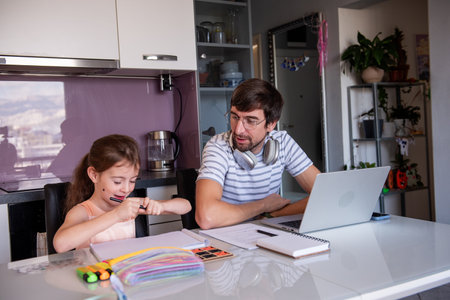 Father is working from home on a laptop with headphones around his neck, while his daughter is sitting next to him and drawing with colored pencilsの写真素材