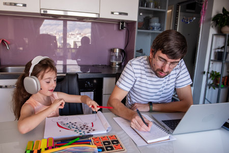Father working from home on a laptop while daughter sits beside him, drawing a rainbow with colored pencils and enjoying music through headphones at the kitchen tableの写真素材