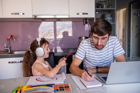 Father is working from home on his laptop, taking notes and taking care of his daughter who is drawing and wearing headphones in their kitchenの写真素材