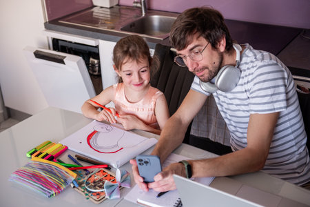 Father with headphones around his neck taking picture of his daughter's drawing of a rainbow with smartphone sitting at kitchen table, open dishwasher in backgroundの写真素材