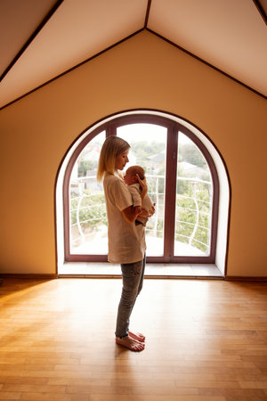Young mother cradling her newborn baby in a sunlit room, standing beside a large arched window, creating a serene and intimate atmosphere filled with love and warmthの写真素材