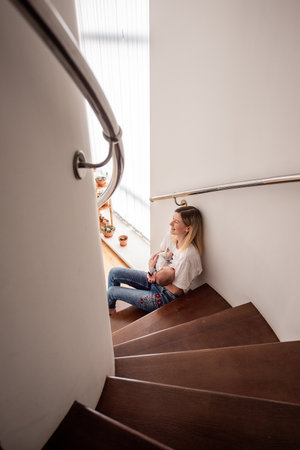 Young mother breastfeeding her newborn baby while sitting comfortably on wooden stairs at home, savoring the joys of motherhood and nurturing moments filled with love and careの写真素材