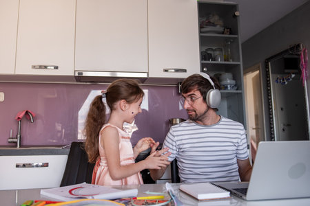 Young father wearing headphones, working on his laptop in the kitchen while his little daughter interrupts with playful chatter and laughter, creating a lively family atmosphereの写真素材