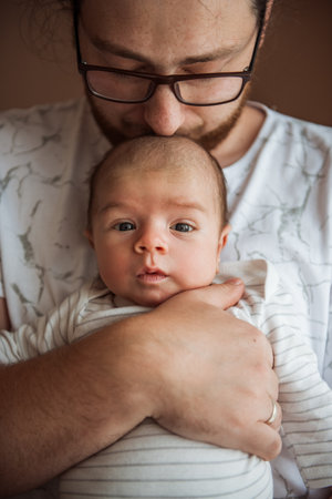 Father wearing glasses kissing his newborn baby on the head while holding him close in a tender embrace, radiating love and care in a warm, cozy moment of connectionの写真素材