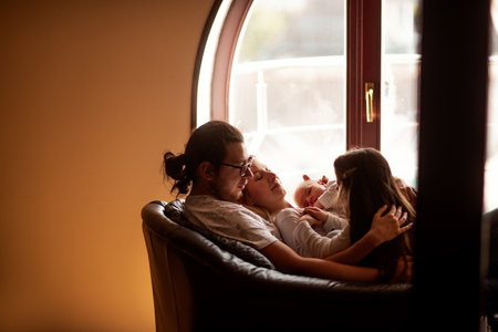 Parents and children are cuddling on a comfortable armchair near a window, enjoying a quiet moment of togetherness and creating a warm family atmosphereの写真素材