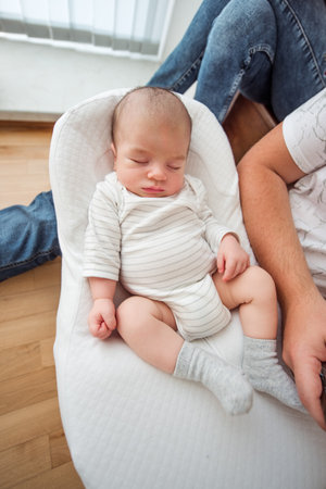 Adorable newborn baby sleeping peacefully in a podster, a safe and comfortable place for infants, with father's legs visible in the background, providing a sense of security and loveの写真素材