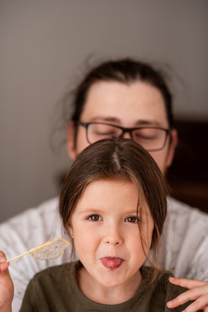 Young girl playfully sticks out her tongue while holding a lollipop. An adult with glasses is in the background, creating a joyful and relaxed atmosphere, emphasizing family fun and bondingの写真素材