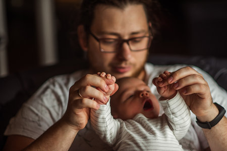 Father lovingly holds his newborn baby's hands, gently lifting them while the baby yawns. The intimate moment captures the warmth and connection of early parenthood in a cozy home settingの写真素材