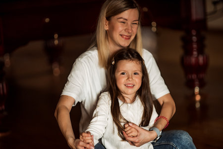 Mother and daughter sitting and holding hands, both wearing white shirts and smiling in a dimly lit room with a wooden floor and some red columns in the backgroundの写真素材