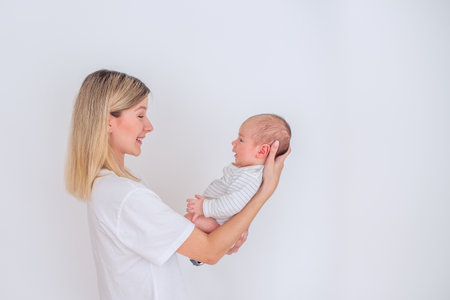 Happy mother holding her newborn baby in her arms, smiling and looking at him, standing in front of a white wall, wearing casual clothes, enjoying motherhoodの写真素材