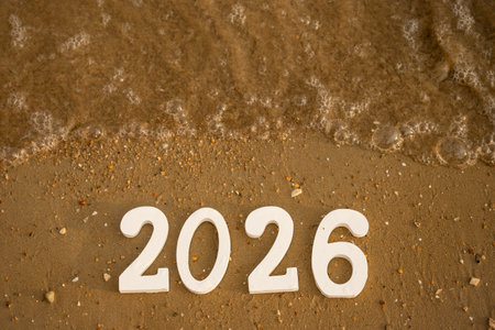 White numbers 2026 on wet sand on a beach, with an incoming wave gently washing them away,...