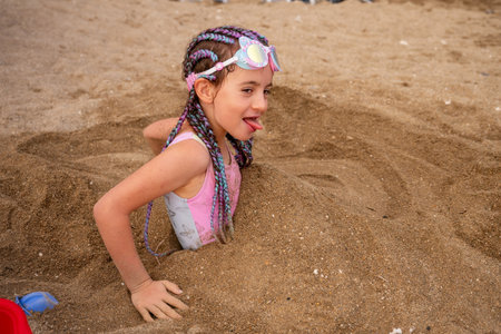 Young girl with braided hair and swimming goggles, playfully buried in sand, sticking out her tongue while enjoying a fun day at the beach during summer vacationの写真素材