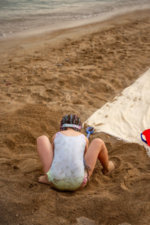 Little girl playing with sand on the beach near the sea, wearing a swimsuit and building sand castles, she is squatting on the beach next to a white towel and a red bucketの写真素材