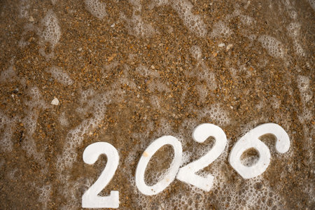 White numbers 2026, partially covered by sea foam, rest on a sandy beach, embodying the spirit of...