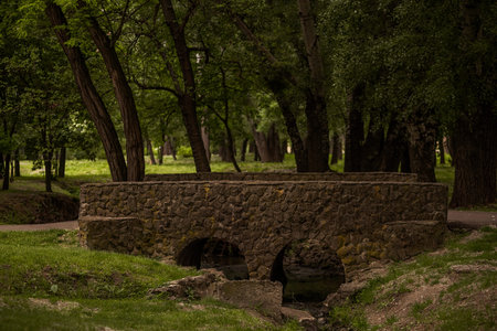 Small stone bridge featuring two graceful arches spans a serene stream in a lush green park,...