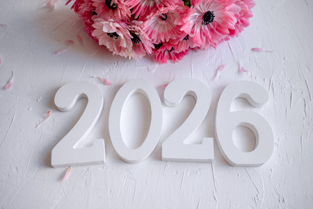 Wooden numbers forming 2026 on a textured white surface with a soft bouquet of pink anemones and...