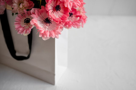 Pink anemone flowers providing a vibrant pop of color, arranged as a luxurious bouquet inside a white paper shopping bag with black handles, symbolizing gifting and celebrationの写真素材