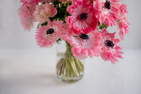 Delicate pink anemone bouquet with dark centers, fresh stems arranged in a clear glass vase on a white table, soft natural light highlighting petals and gentle spring eleganceの写真素材