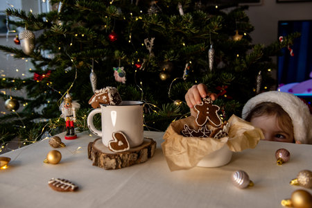 Young child's hand reaching for a gingerbread cookie from a bowl of holiday treats next to a mug of...
