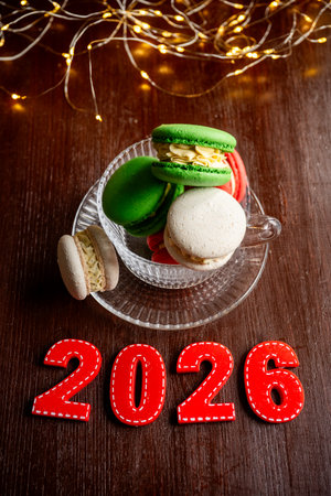 Colorful macarons sitting in a glass teacup next to red 2026 numbers, illuminated by warm string lights on a dark wooden background, symbolizing holiday joy and upcoming celebrationsの写真素材