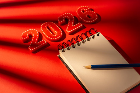 Red numbers 2026 are next to an open blank notebook and a blue pencil ready for writing down New year resolutions, future goals, and personal development plans on a vibrant red backgroundの写真素材